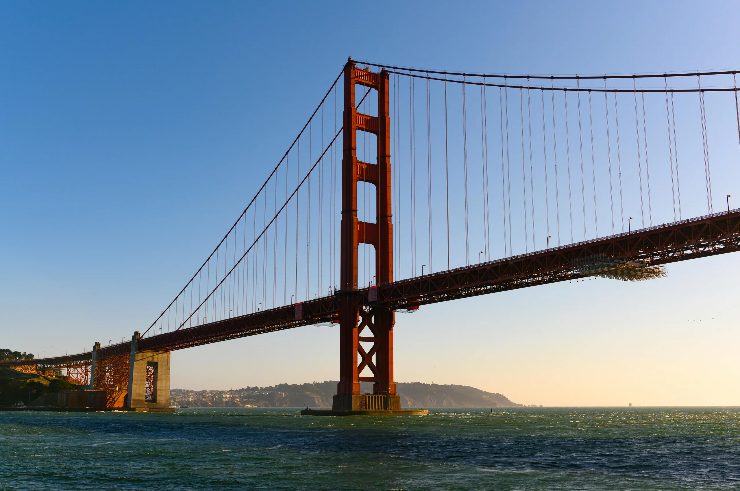 A low-angle view of the iconic orange Golden Gate Bridge spanning across calm water under a clear blue sky.