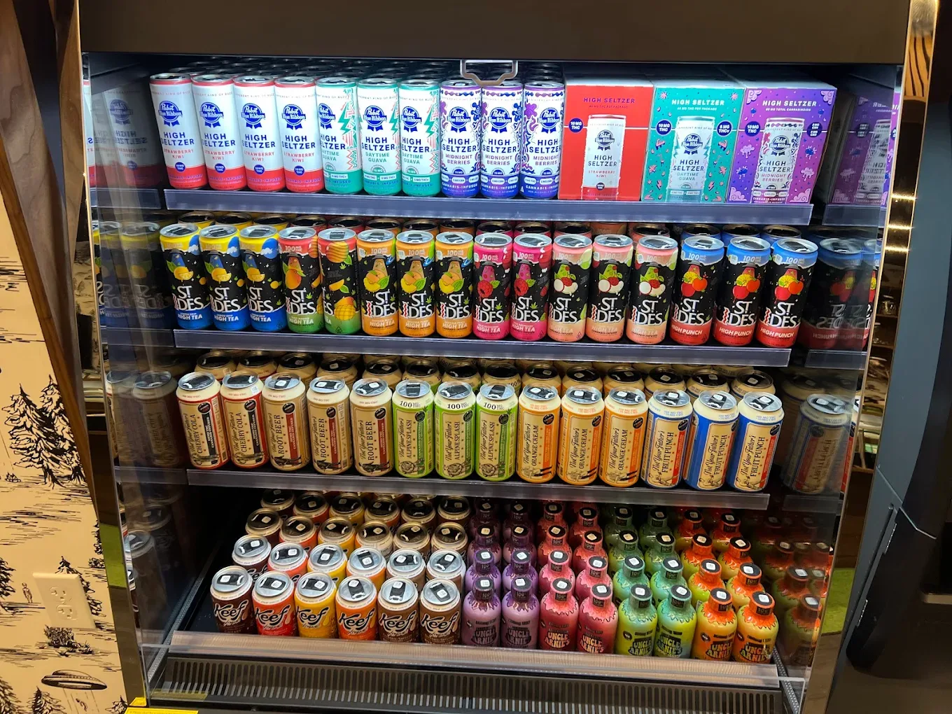 A refrigerated display case is fully stocked with various brands of canned beverages, including Pabst Blue Ribbon High Seltzer, St. Ides, and other flavored drinks.